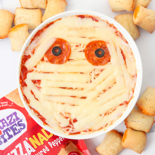 Cheesy mummy dip in a bowl surrounded by Brazi Bites Pizza'nadas